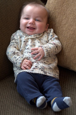 A mostly-bald white baby sits supported in the corner of a brown couch. He's got a big smile and is wearing a speckled ivory-brown sweater, blue jeans, and blue-tan striped socks. 